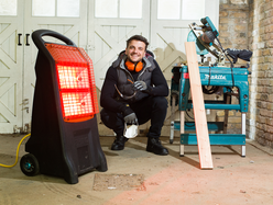 A portable infrared heater glowing red in a workshop, positioned near a worker and woodworking tools on the floor.