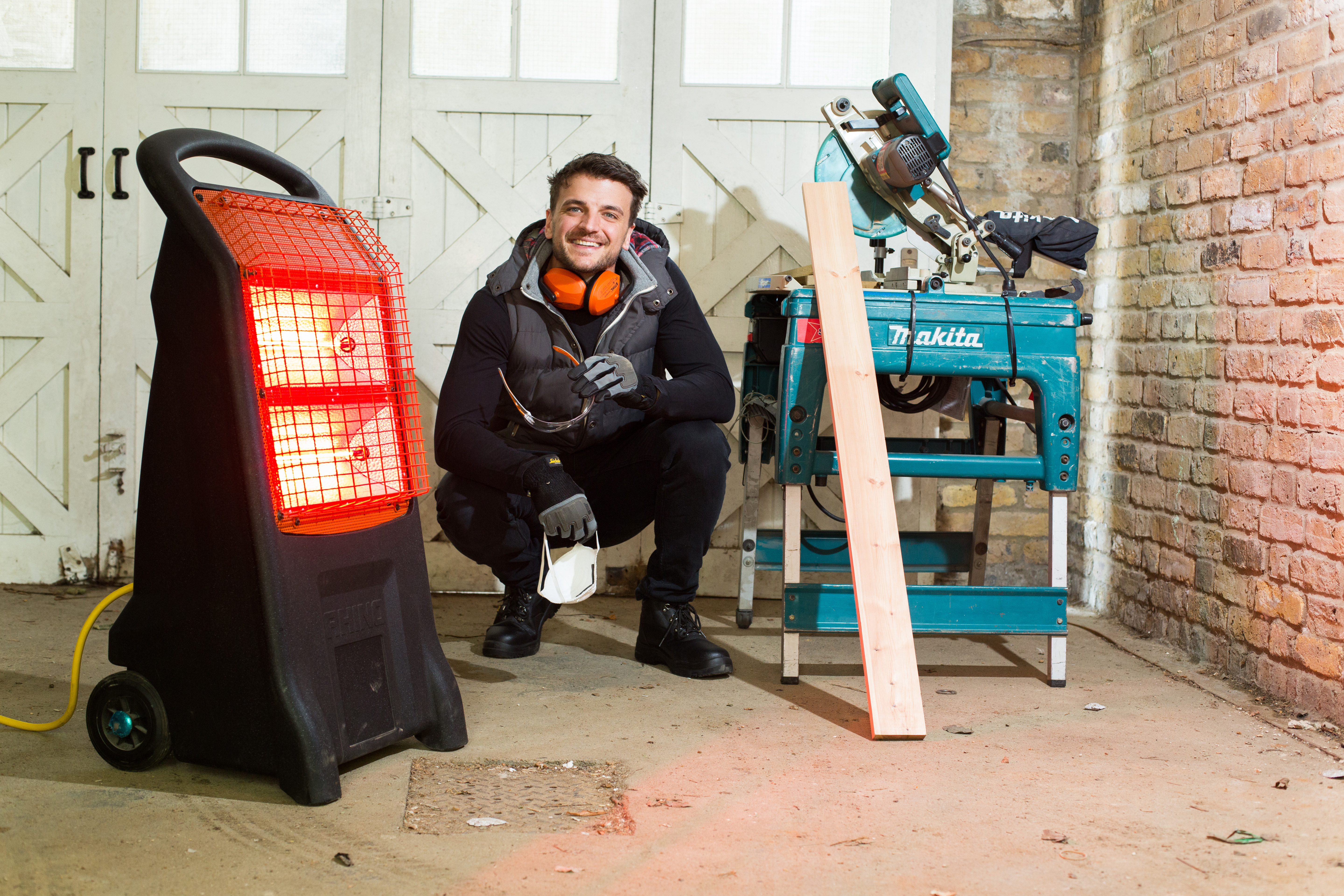 A portable infrared heater glowing red in a workshop, positioned near a worker and woodworking tools on the floor.