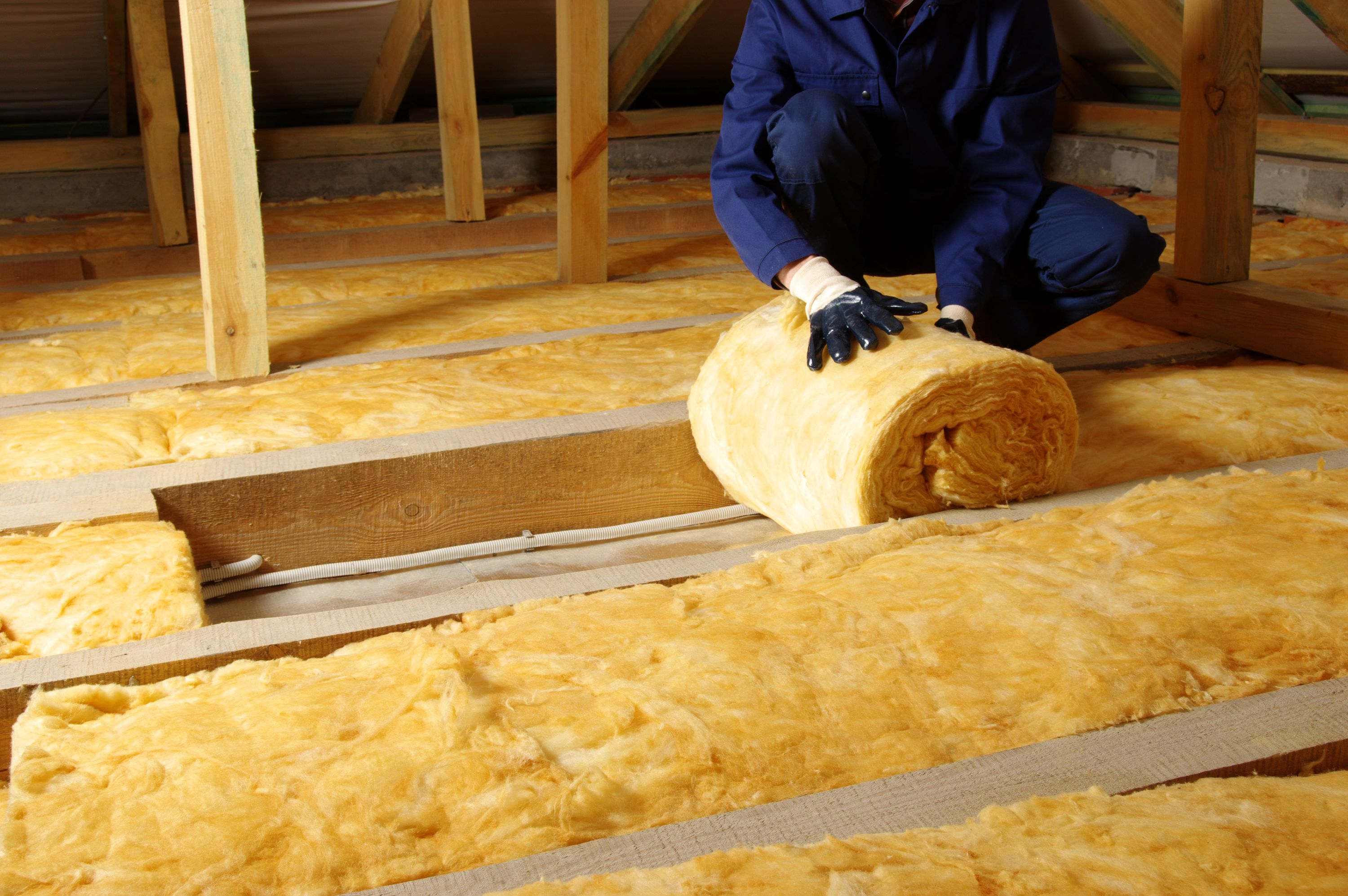 Worker laying rolls of yellow insulation material across attic floor joists.