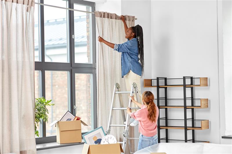 Two people working together on a step ladder to hang curtains in a bright room with moving boxes and shelving.