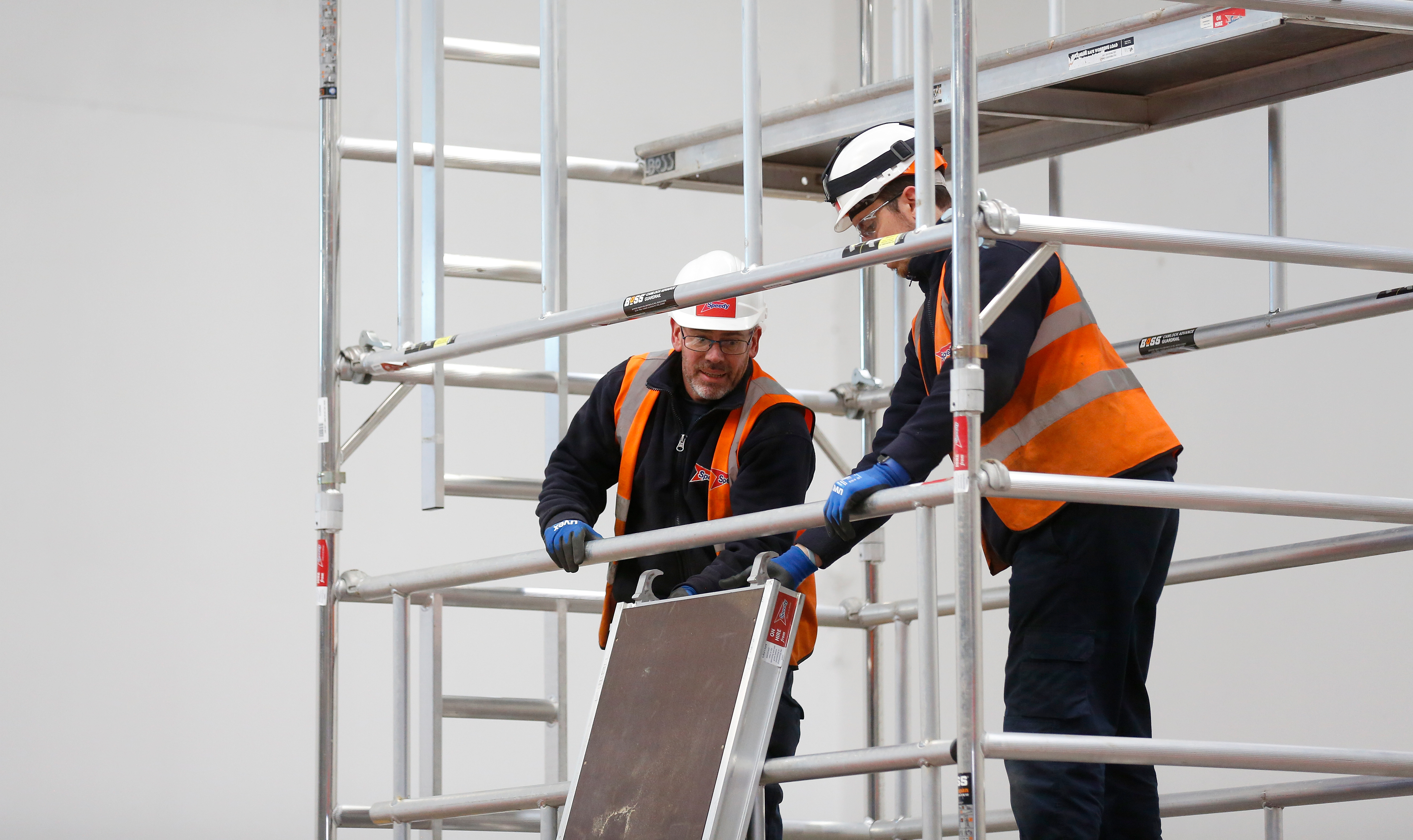 Two workers wearing safety helmets, gloves, and orange high-visibility vests installing a platform on an aluminum scaffold structure indoors