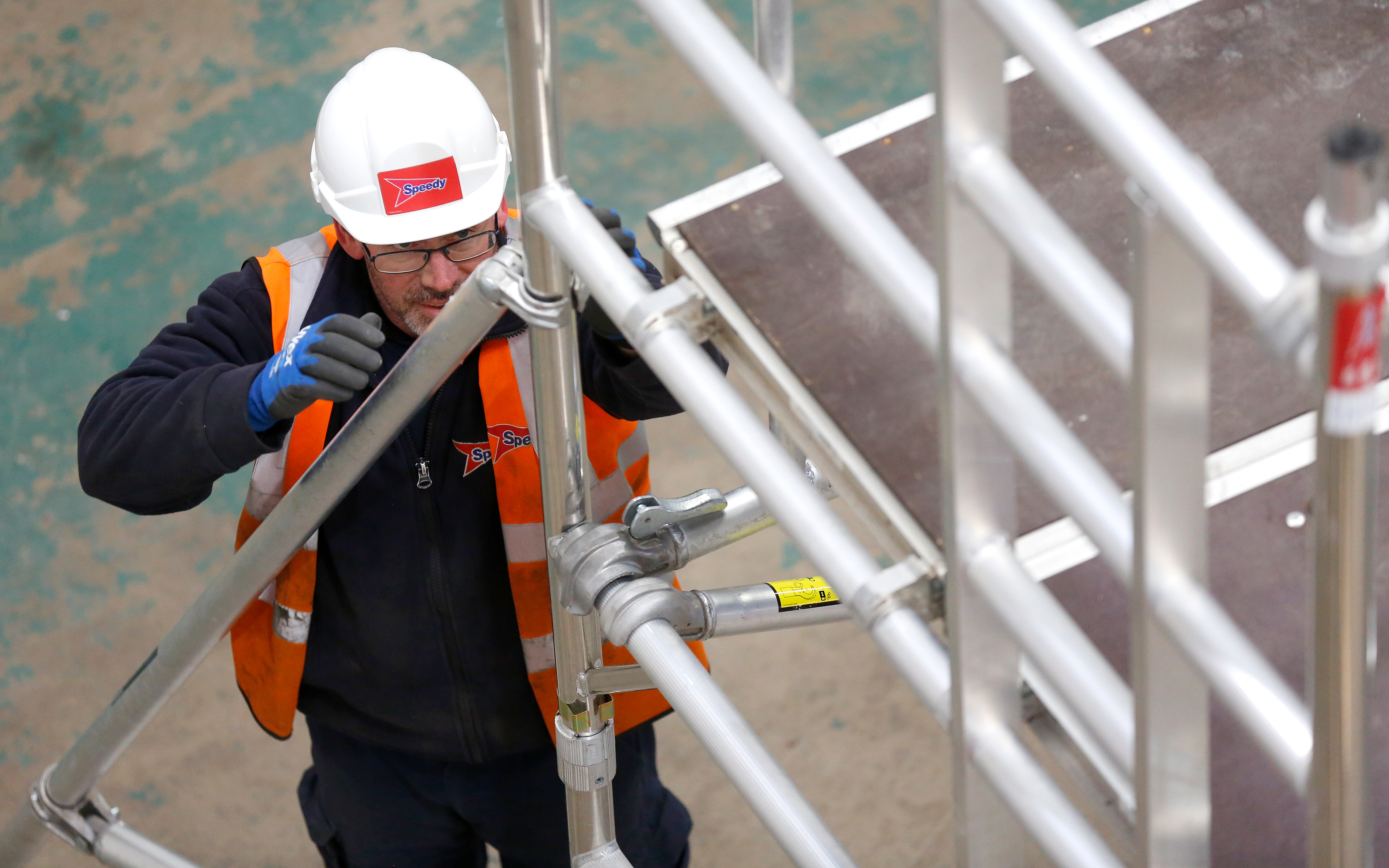 A worker wearing a white safety helmet and an orange high-visibility vest is assembling an aluminium scaffold stabiliser indoors.