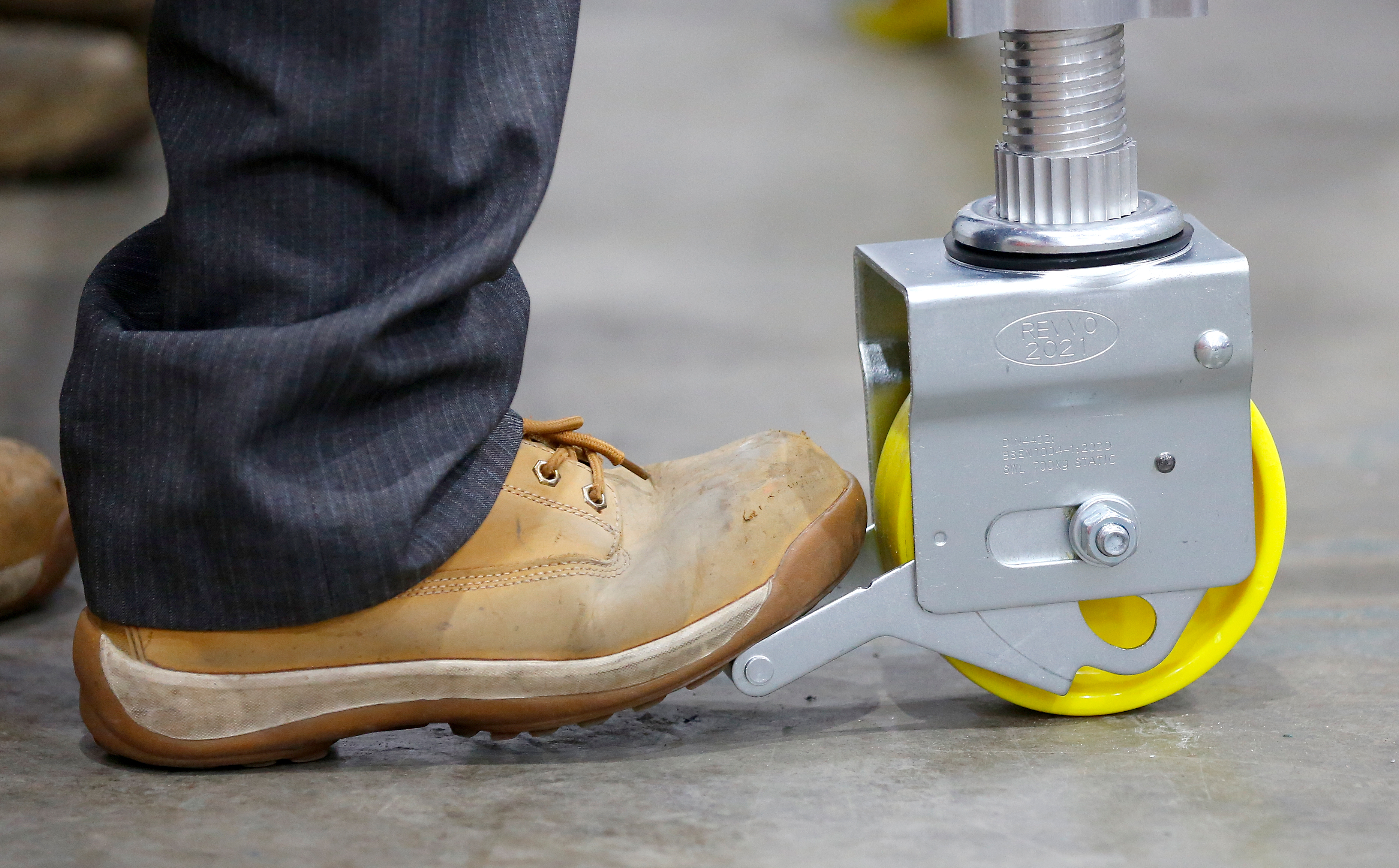 Close-up of a person&rsquo;s boot pressing the brake lever on a yellow caster wheel attached to a mobile scaffold frame on a concrete floor.