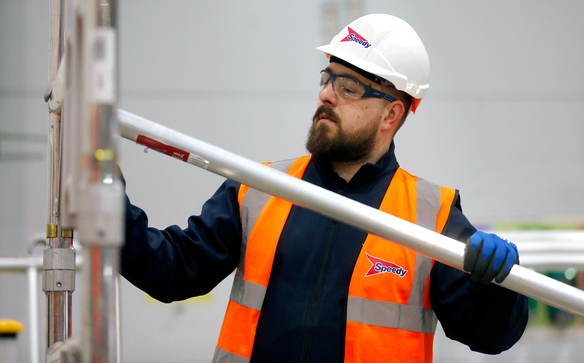 Worker wearing Speedy-branded safety gear assembling aluminum scaffold frame indoors.