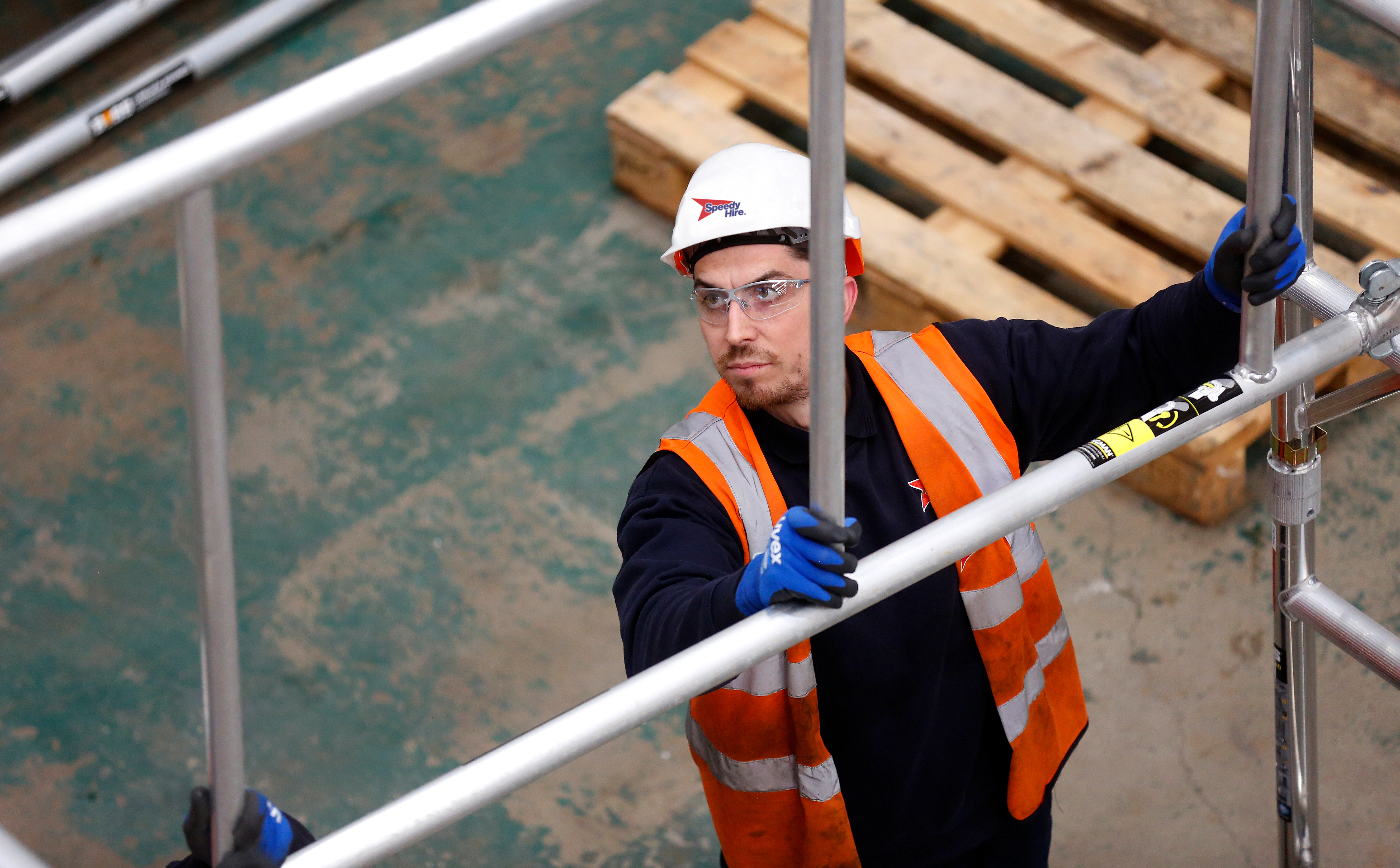 Worker wearing Speedy-branded safety gear assembling aluminum scaffold frame indoors.