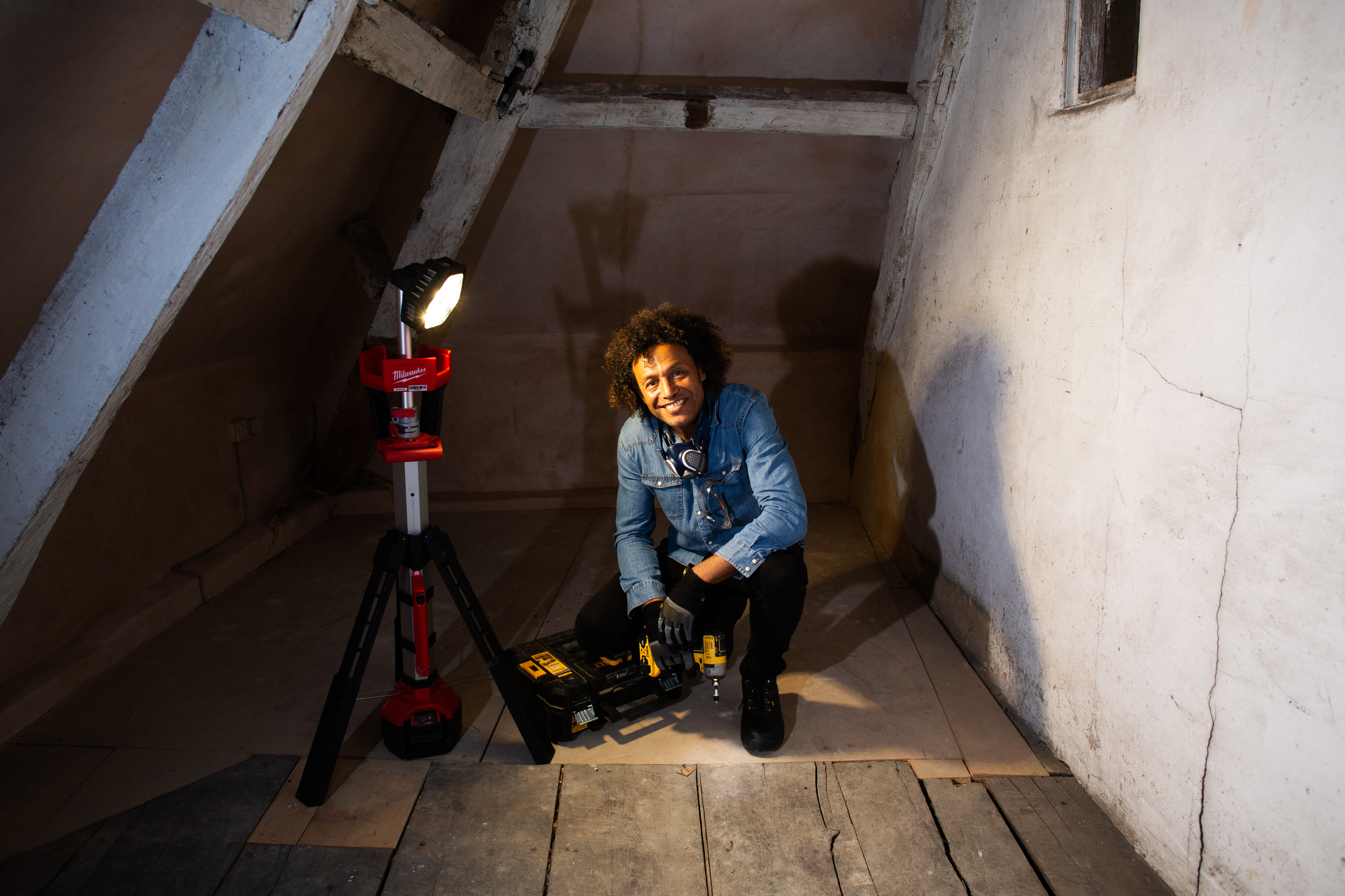 A person kneeling in an attic while organising power tools beside a Milwaukee battery light